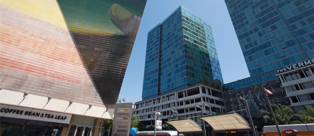 Low angle view of modern high rise buildings in a city setting under a clear blue sky. On the left is a large angular building with a slanted fa&ccedil;ade covered by a large digital style display showing abstract gradient colors and horizontal lines. At street level beneath it is a storefront with signage reading COFFEE BEAN & TEA LEAF. In the center and right are tall glass office towers with reflective windows. The building on the far right displays signage near the top reading THE VERMONT. In the foreground are street elements including light poles, a bus stop canopy, a city bus partially visible, palm trees, and a sign listing nearby businesses.
