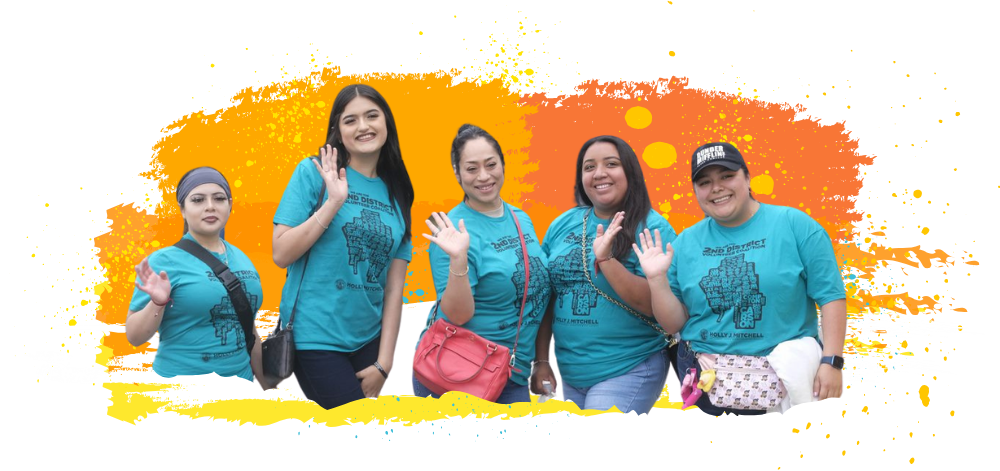 Five people stand side by side smiling and waving, wearing matching teal shirts with text that reads 2ND DISTRICT and HOLLY J. MITCHELL beneath a graphic design. One person wears a black hat with text that reads SUPER VOLUNTEER. The group stands in front of a bright orange and yellow paint splash graphic on a black background.